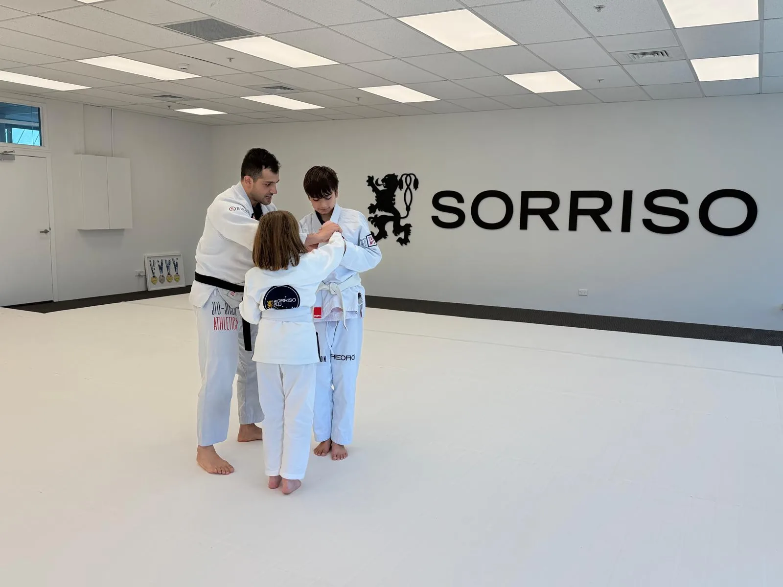 A black belt coach guides two children through a grip drill on the white mats at Sorriso BJJ Botany